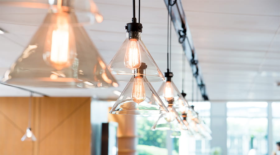 Primo piano di lampade a sospensione in vetro con lampadine stile edison accese, installate in un ambiente moderno con sfondo sfocato di persone sedute a un tavolo.