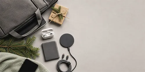 Idee regalo di Natale tecnologiche per colleghi: auricolari wireless, power bank e caricabatterie wireless accanto a un pacchetto regalo.